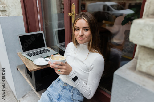 Canvas Print young woman holding cup of cappuccino while sitting in outdoor cafe next to laptop with blank screen in Vienna