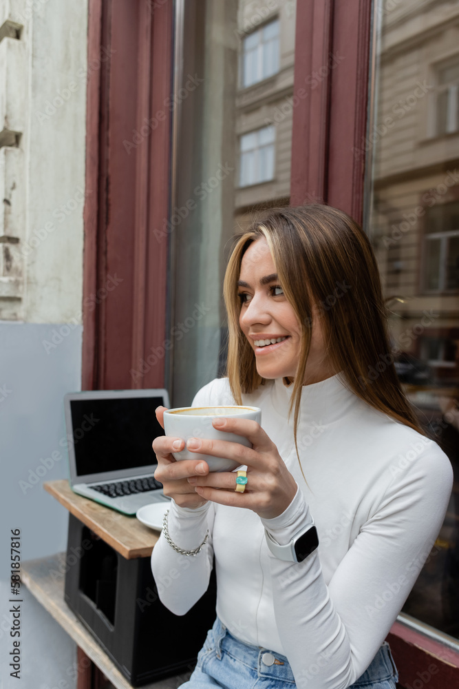 pretty woman holding cup of coffee while sitting in outdoor cafe next to laptop with blank screen in Vienna. pretty woman holding cup of coffee while sitting in outdoor cafe next to laptop with blank screen in Vienna.
