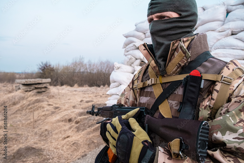 Soldier with weapon in military uniform stands next to the barricades made from sandbags and anti-tank hedgehog barriers. Military man on the roadblock. Combatant in full ammunition. Concept of war Soldier with weapon in military uniform stands next to the barricades made from sandbags and anti-tank hedgehog barriers. Military man on the roadblock. Combatant in full ammunition. Concept of war