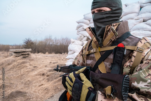 Soldier with weapon in military uniform stands next to the barricades made from sandbags and anti-tank hedgehog barriers. Military man on the roadblock. Combatant in full ammunition. Concept of war
