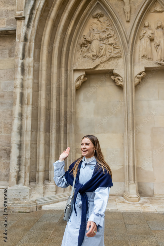 cheerful young woman with scarf on top of blue trench coat waving hand near historical building in Vienna.