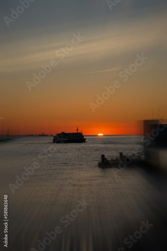 sunset with a silhouetted boat sailing just out of port along its journey against a vivid colorful sunset an orange and yellow color filled sky with motion blur. 