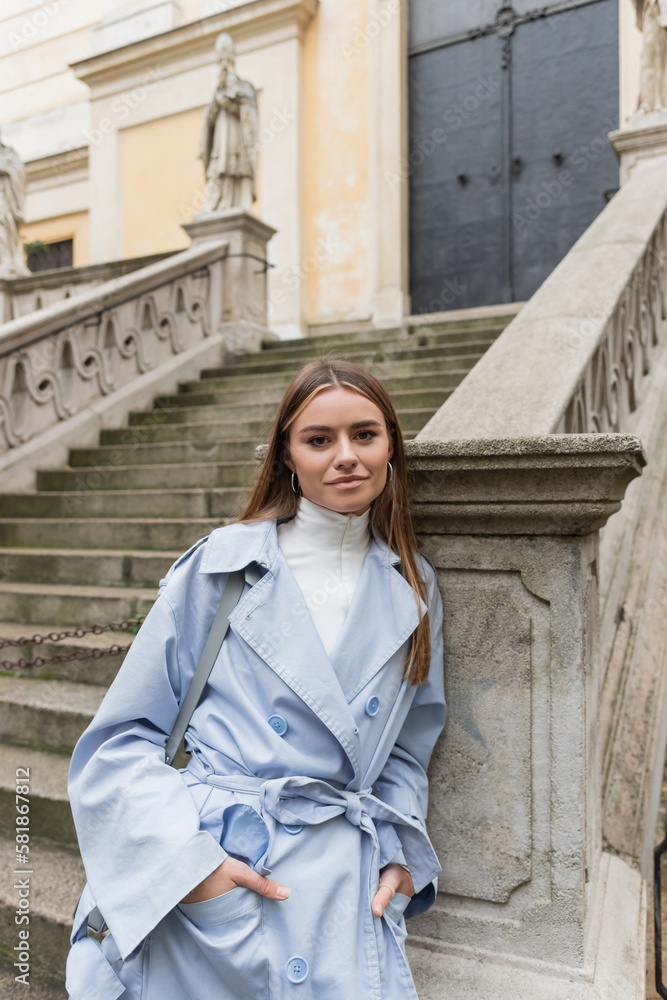 charming woman in blue trench coat standing with hands in pockets near ancient stairs on street in in Vienna.