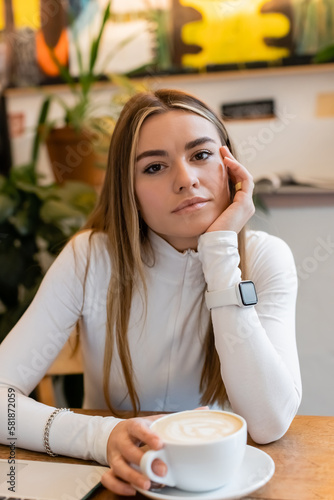 young woman in turtleneck with smart watch sitting next to cup of cappuccino in cafe.