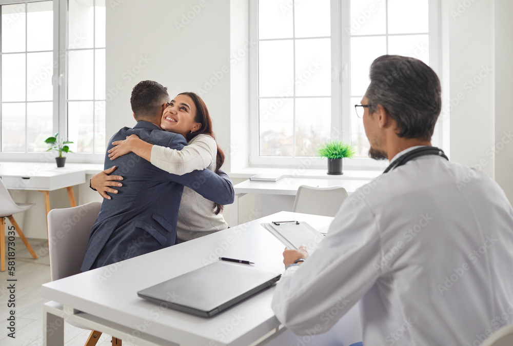 Fototapeta premium Young married couple are happy, they will soon become parents. Good news from friendly doctor at meeting in office of hospital or clinic. Joyful husband and wife at pregnancy planning consultation.