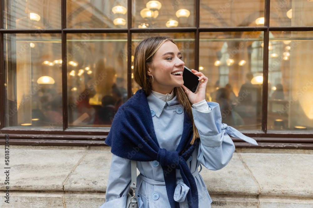 Fototapeta premium cheerful woman with scarf on blue trench coat talking on smartphone on street in Vienna.