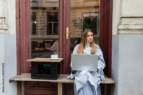 young woman in blue trench coat using laptop while sitting next to cup of coffee in outdoor cafe in Vienna.