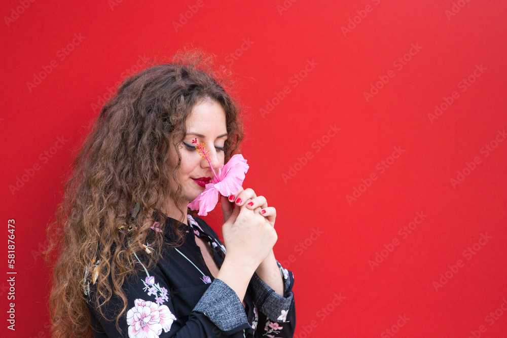 Beautiful woman, young and blonde with curly hair and blue eyes is smelling a pink flower of Hawaiian scientific name inhibisco. The woman holds the flower in her hands on red background. Beautiful woman, young and blonde with curly hair and blue eyes is smelling a pink flower of Hawaiian scientific name inhibisco. The woman holds the flower in her hands on red background.