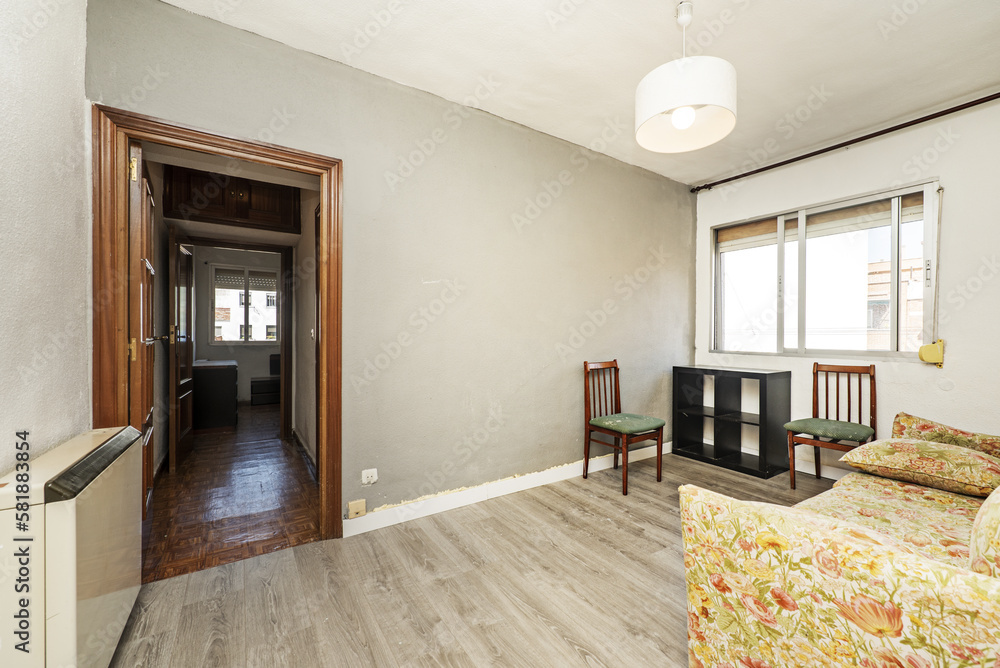 Fototapeta premium Living room furnished with a solitary sofa in a patterned fabric of dubious taste, sapele wooden doors and gray wooden floors, poorly painted walls and a ceiling lamp