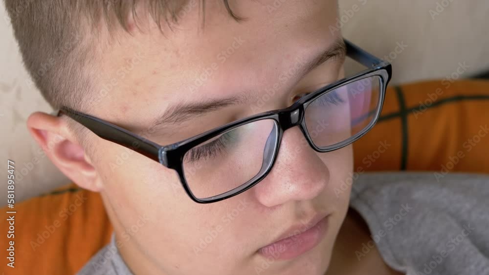 Close-up Face of a Thinking Serious Boy with Glasses, with Full Lips. Face portrait of a tired child, with long eyelashes, looking at a TV in bed. Body part. Emotions. Problems, poor eyesight. Macro. Close-up Face of a Thinking Serious Boy with Glasses, with Full Lips. Face portrait of a tired child, with long eyelashes, looking at a TV in bed. Body part. Emotions. Problems, poor eyesight. Macro.