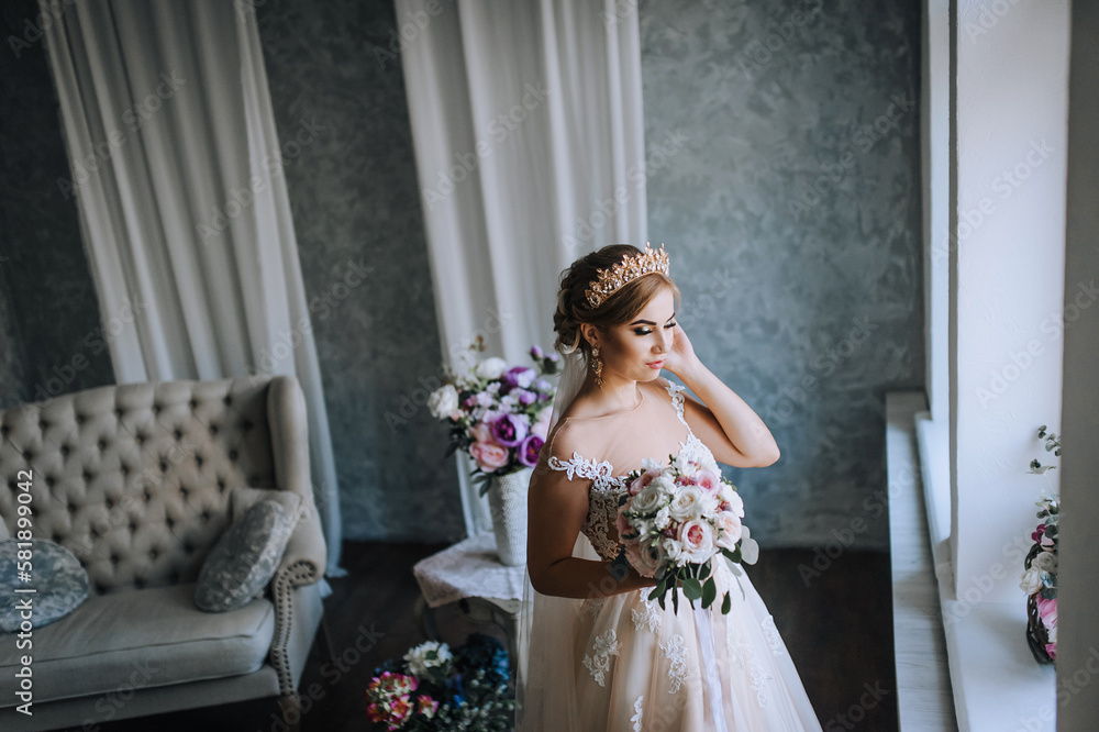 A beautiful young blonde model bride in a white lace dress with a crown, a diadem on her head stands in the studio, indoors with a bouquet in her hands near the window. Wedding photography, portrait. A beautiful young blonde model bride in a white lace dress with a crown, a diadem on her head stands in the studio, indoors with a bouquet in her hands near the window. Wedding photography, portrait.
