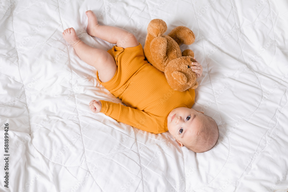 cute baby in a yellow bodysuit is lying with a teddy bear in bed. Top