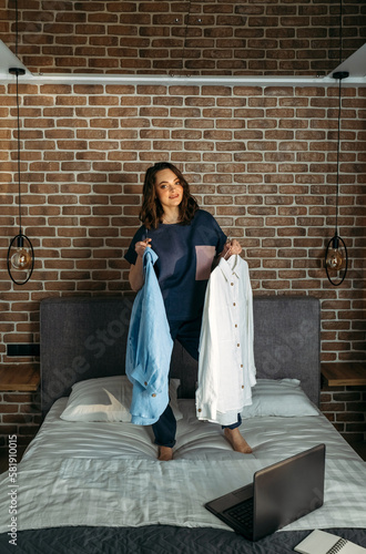 A beautiful woman in designer clothes stands on the bed and holds clothes bought online in her hands.
