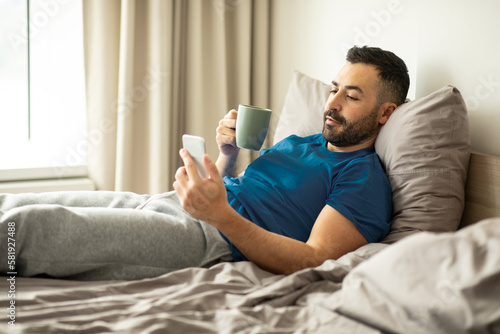 Tapeta European man using cellphone and drinking coffee, holding cup and browsing inter