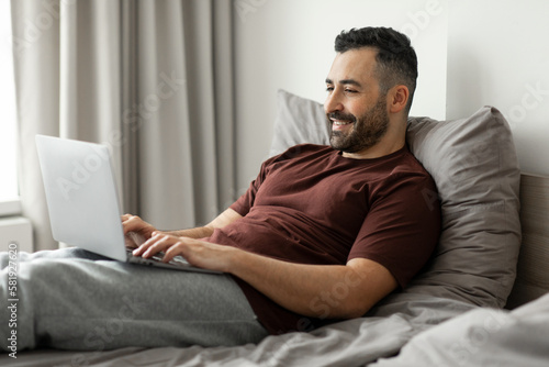 Excited middle aged man using laptop while sitting in bed at home, looking at screen and typing on computer keyboard, enjoying remote job and freelancing, free space