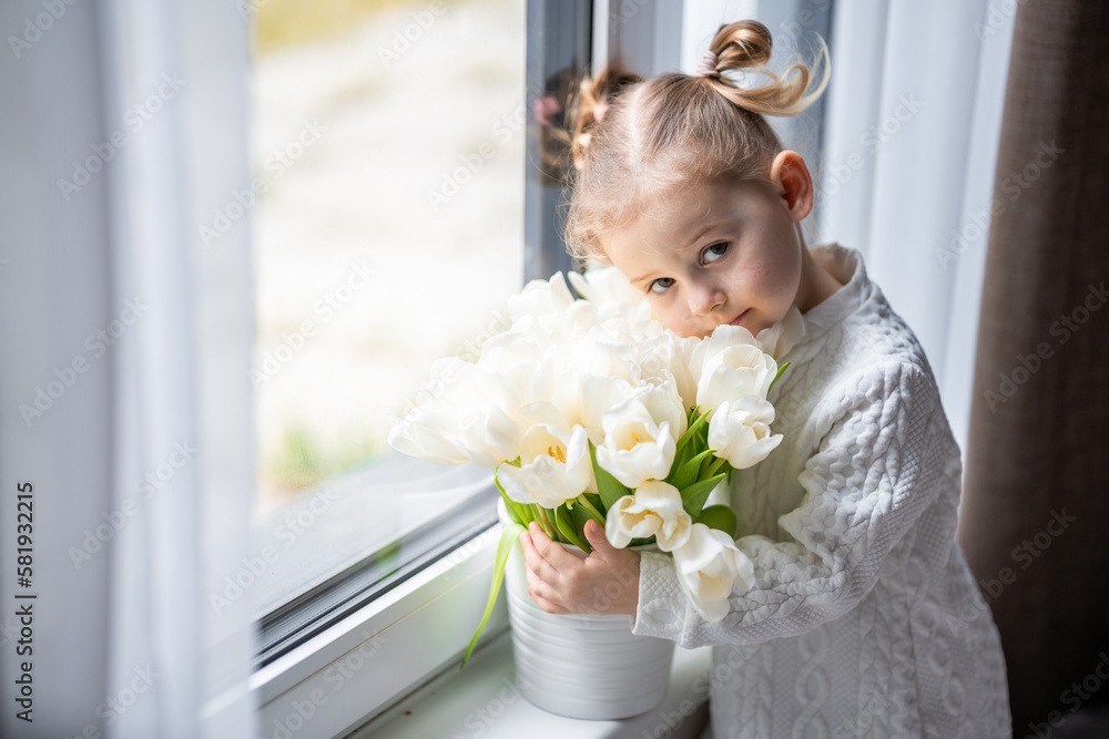 Little girl sitting by window with tulip flowers bouquet. Happy child, indoors. Mother's day, valentine's day or birthday concept. Little girl sitting by window with tulip flowers bouquet. Happy child, indoors. Mother's day, valentine's day or birthday concept.