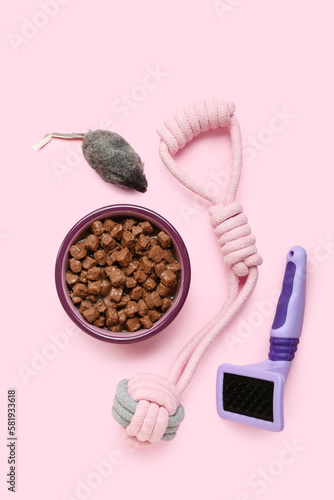 Bowl of wet pet food  grooming brush and toys on pink background