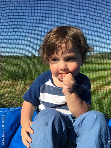 cute smile curly baby boy holding his finger in his mouth sitting on a trampoline