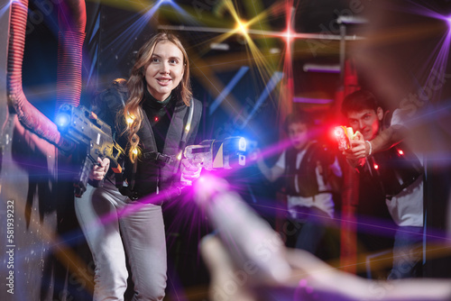Joyous girl with laser pistol playing laser tag with friends on dark labyrinth