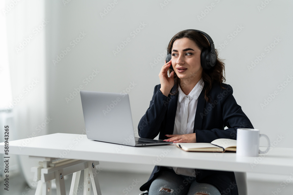 An attractive girl is sitting at a desk near a laptop in headphones An attractive girl is sitting at a desk near a laptop in headphones