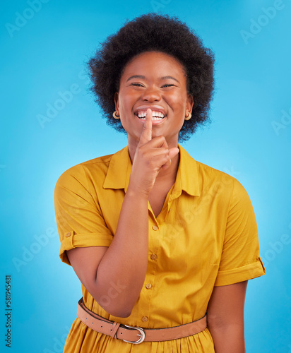 Portrait, smile and secret with a black woman in studio on a blue background standing finger on lips. Gossip, whisper and mystery withan attractive young female making a hand gesture emoji of privacy photo