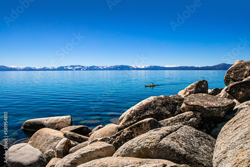 Secret Cove panoramic view of Lake Tahoe on clear day with Kayaker in the water and snow-covered mountains of the Sierra Nevada in the background