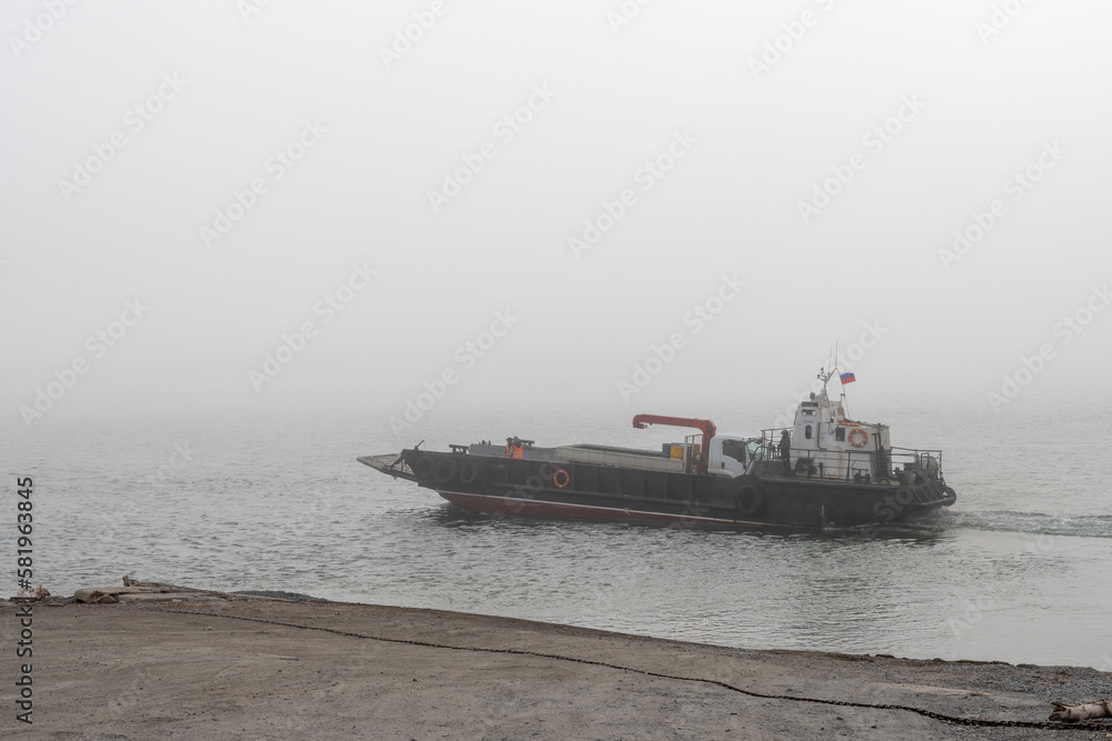 A barge transports a truck across a river. Poor visibility in fog. Passenger and automobile water transportation on a barge. Yana river, Magadan region, Siberia, Far East of Russia. Foggy weather. A barge transports a truck across a river. Poor visibility in fog. Passenger and automobile water transportation on a barge. Yana river, Magadan region, Siberia, Far East of Russia. Foggy weather.