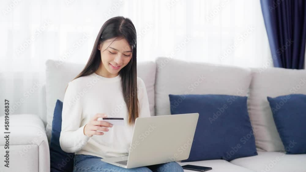 Young asian woman sitting on sofa in living room, makes online banking payments through the internet from bank card on computer laptop. Shopping online on notebook with credit card Young asian woman sitting on sofa in living room, makes online banking payments through the internet from bank card on computer laptop. Shopping online on notebook with credit card