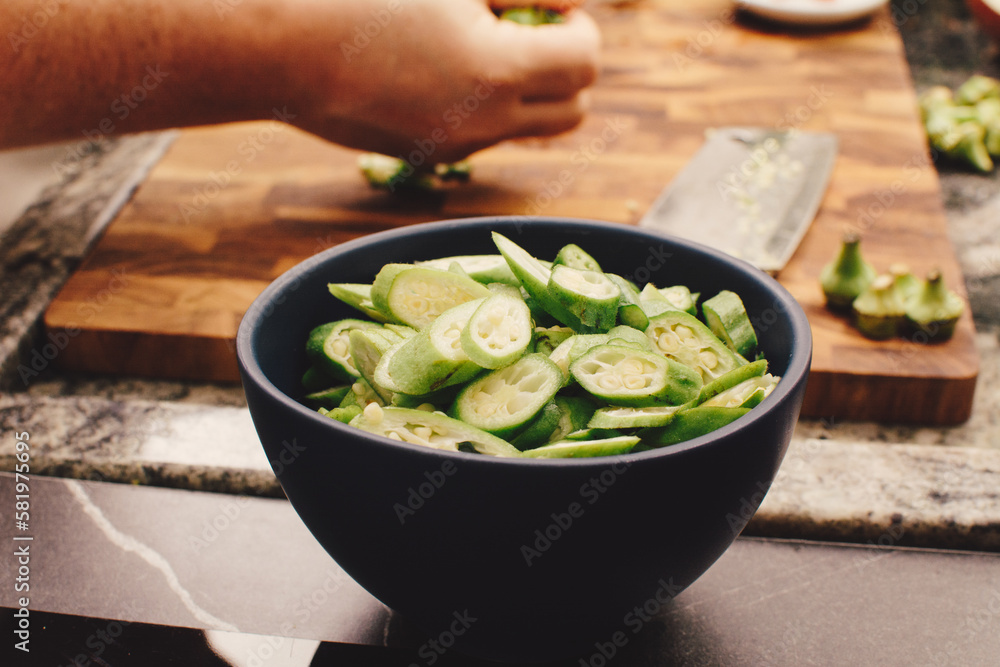 Sliced okra in blue ceramic bowl with female hands cutting more on wooden cutting board in background Sliced okra in blue ceramic bowl with female hands cutting more on wooden cutting board in background