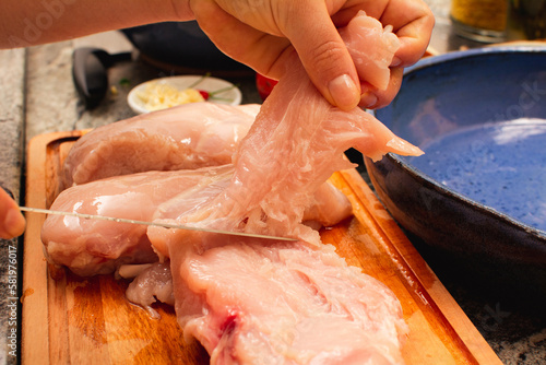 Woman slicing raw chicken breast fillets on bamboo cutting board in kitchen (close-up)