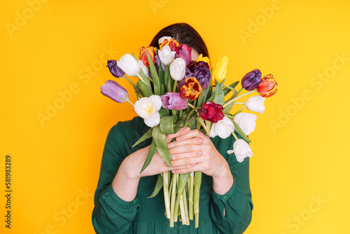 Lady in green dress covered her face with bouquet of tulips against yellow background.