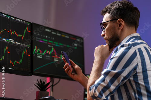 Night portrait of serious handsome trader holding mobile phone, using laptop computer working online with crypto chart sitting in modern office. Successful business, online trading concept 