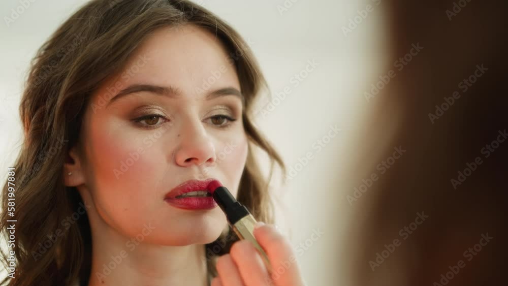 Focused young woman paints lips with glamorous red lipstick looking in mirror. Female person does bright makeup to go to birthday party closeup Focused young woman paints lips with glamorous red lipstick looking in mirror. Female person does bright makeup to go to birthday party closeup