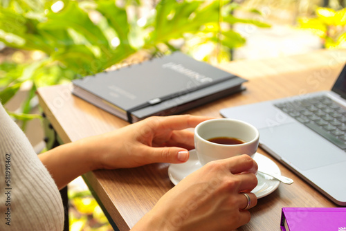 Mãos femininas segurando uma xicara de café branca em uma mesa de trabalho arrumada e organizada com um notebook e uma agenda com flores ao fundo photo