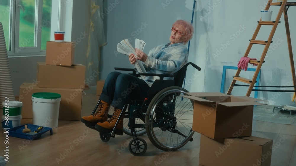 An elderly disabled woman in a wheelchair looks through the palette with colors. A granny plans repairs and chooses a paint color. Room with window, ladder, cardboard boxes, wallpaper, paint.