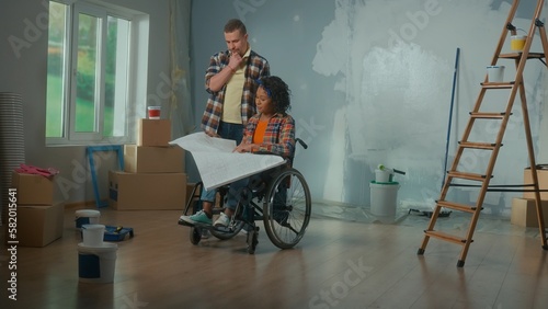A white man and an African American disabled woman in a wheelchair are looking at an apartment project sheet. A young couple is planning a renovation in room, talking and discussing interior design.