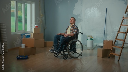 A young man in a wheelchair. A disabled man thinks and plans repairs in the room. Handicapped person on the background of cardboard boxes, cans of paint and a ladder.