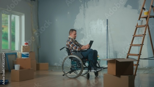 A young man in a wheelchair is planning repairs using a digital tablet. Disabled male browsing interior design ideas on the internet. Handicapped person.