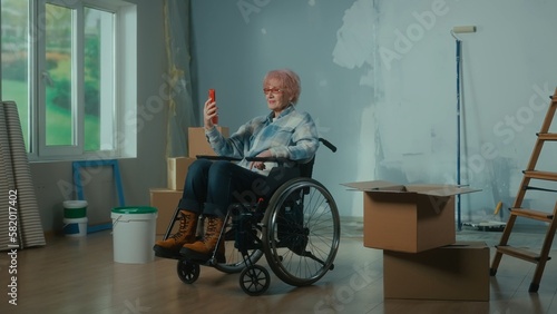 An elderly disabled woman in a wheelchair talks on a video call using a mobile phone. A room with a window, ladder, wallpaper, cardboard boxes.