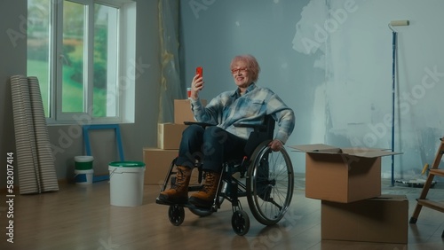 An elderly disabled woman in a wheelchair talks on a video call using a mobile phone. A room with a window, ladder, wallpaper, cardboard boxes.