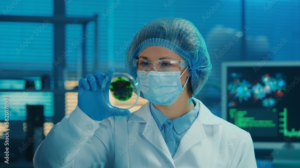 A female scientist examines a petri dish with a spotted green sample. A woman in a white gown, goggles, mask, blue gloves and a bonnet examines the spores of a virus or bacteria. Close up. A female scientist examines a petri dish with a spotted green sample. A woman in a white gown, goggles, mask, blue gloves and a bonnet examines the spores of a virus or bacteria. Close up.