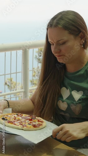 A young woman sitting in a cafe. She sets aside an empty plate and takes a pizza. Sea view. Vertical. Social Media photo