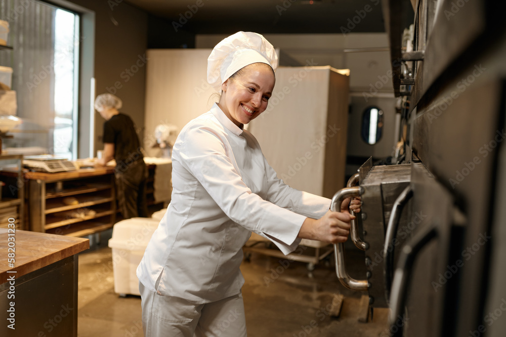 Smiling woman baker opening big industrial oven at modern bakery kitchen