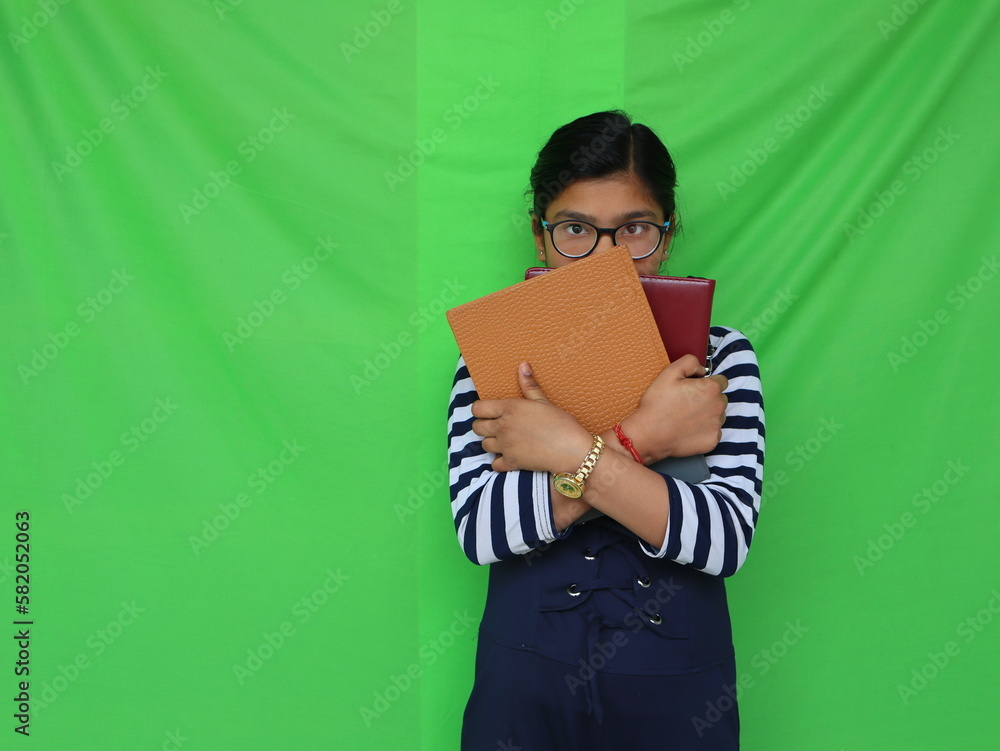 Obraz premium Specs wearing teenage girl holding books in his hand and hiding his face behind books, green background 