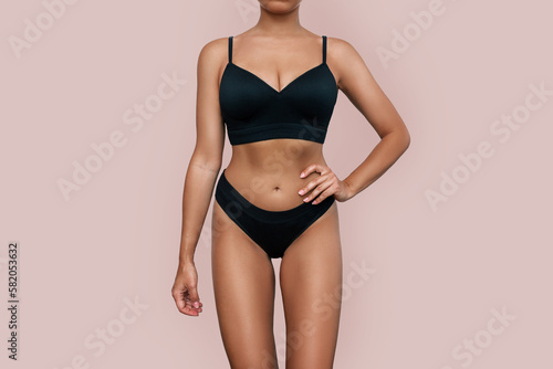 Cropped shot of young tanned fit woman in black underwear demonstrating her slender figure with toned abs isolated on beige background. Result of fitness, diet, healthy lifestyle. Female perfect body
