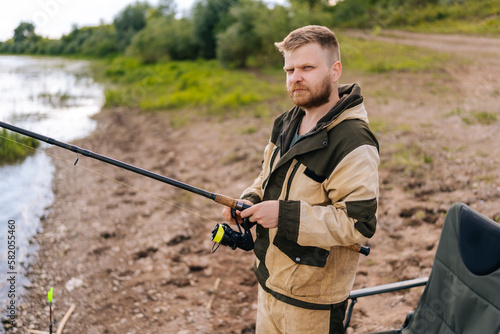 Portrait of bearded fisher man holding spinning casting rod standing on bank waiting for bites on water river at summer day, looking at camera. Concept of lifestyle, leisure activity on nature