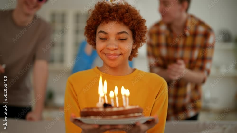 Portrait smiling african american birthday girl with cake in hands looks at burning candles makes a wish. Happy party with friends clapping hands and blowing horns on background. Celebration concept. Portrait smiling african american birthday girl with cake in hands looks at burning candles makes a wish. Happy party with friends clapping hands and blowing horns on background. Celebration concept.