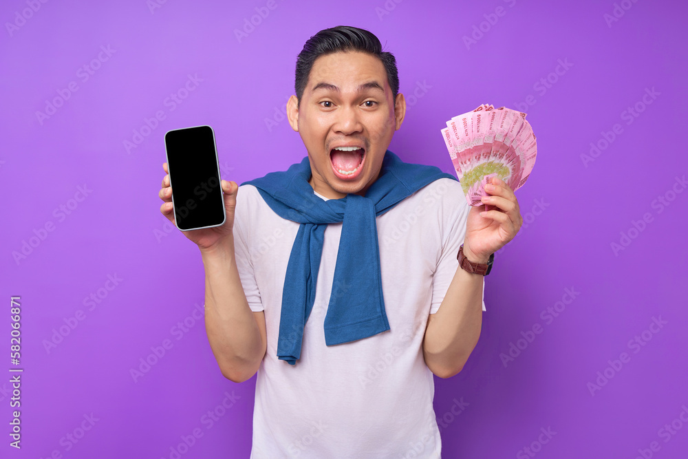 Excited young Asian man in white t-shirt showing blank screen mobile phone and holding money banknotes isolated on purple background Excited young Asian man in white t-shirt showing blank screen mobile phone and holding money banknotes isolated on purple background