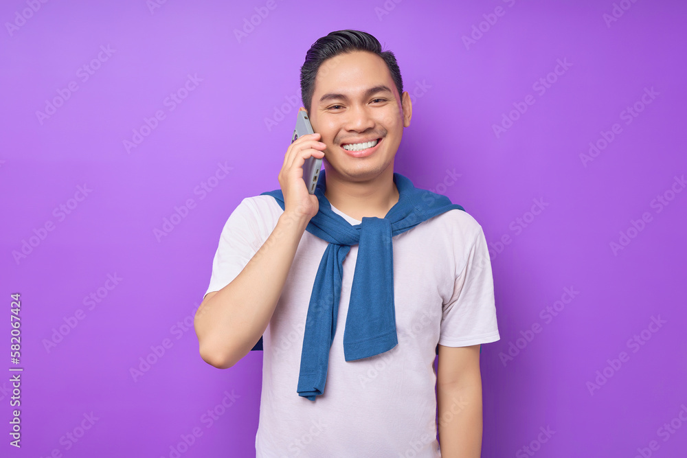 Cheerful young Asian man in white t-shirt talking on mobile phone isolated on purple background