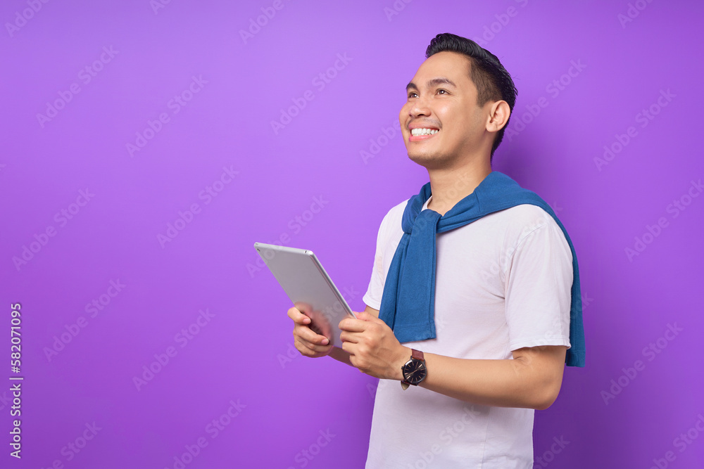 Smiling young Asian man in casual clothes holding digital tablet and looking aside at copy space isolated over purple background